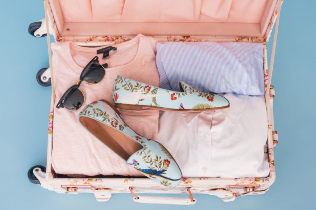 A suitcase packed with light pastel-colored clothing, sunglasses, and a pair of shoes with a flower motif. 