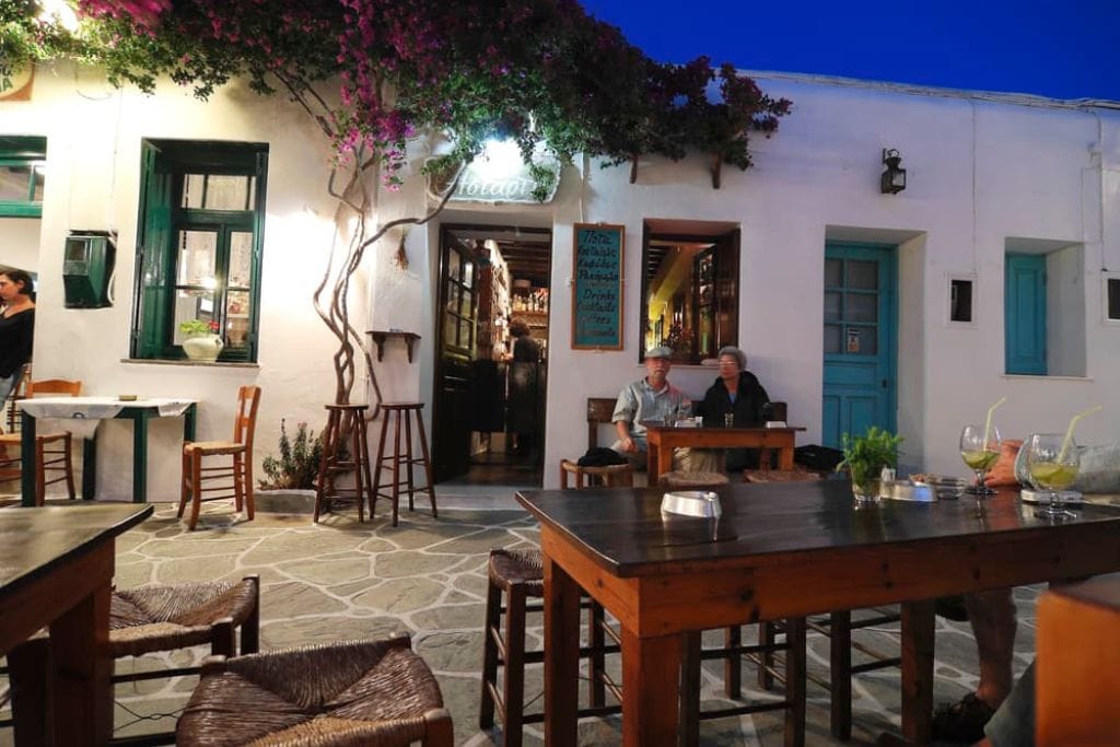An older man and woman sit outside a restaurant on a bench in Greece.