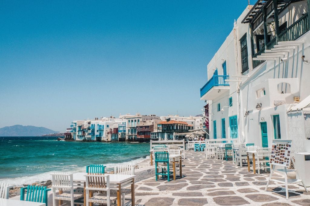 A restaurant with empty tables and chairs sit along the water in Greece. 