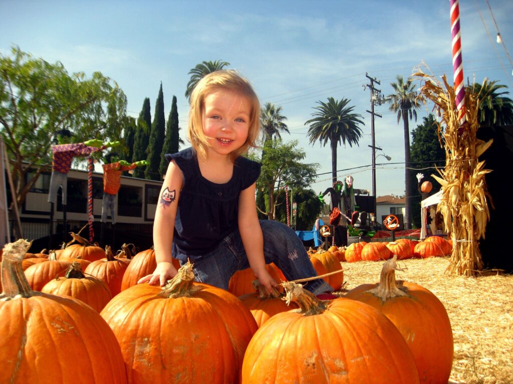 Beautiful little girl trying to pick up a giant orange pumpkin at a pumpkin patch, with hay bails and pumpkins surrounding her.