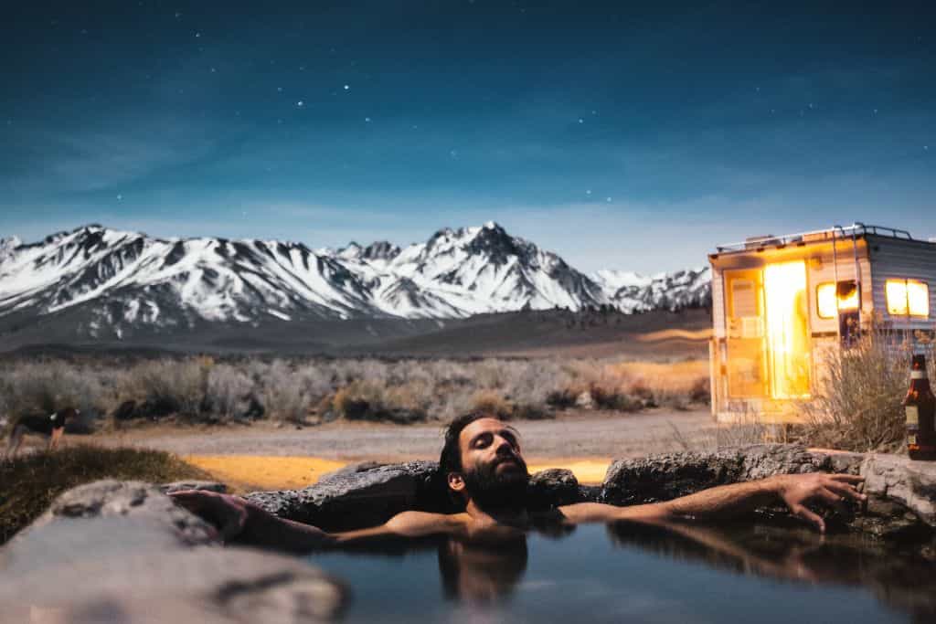 A man with a dark beard soaking in a natural hot spring surrounded by the snow-capped Sierra Mountains.
