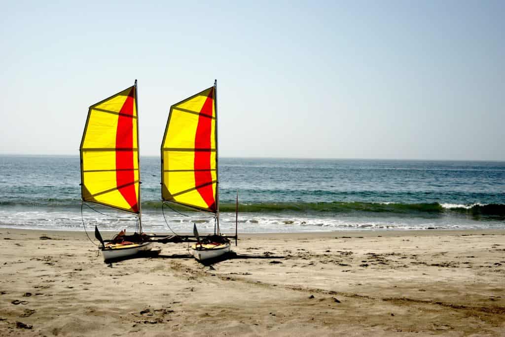 A catamaran with yellow and red sails sit parked on an empty beach with the ocean behind it. Bodega Bay in August.