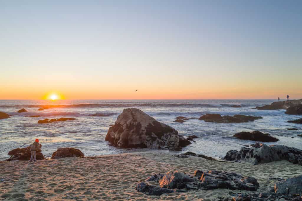 The sun setting over the Pacific Ocean with a beach and rock formations in the foreground. Bodega Bay in December.