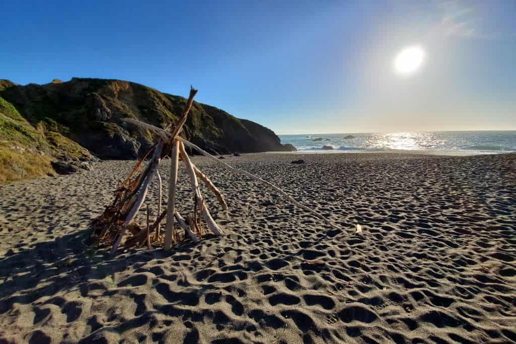 A tent made of driftwood on the beach with the sun and a cliff in the background. 