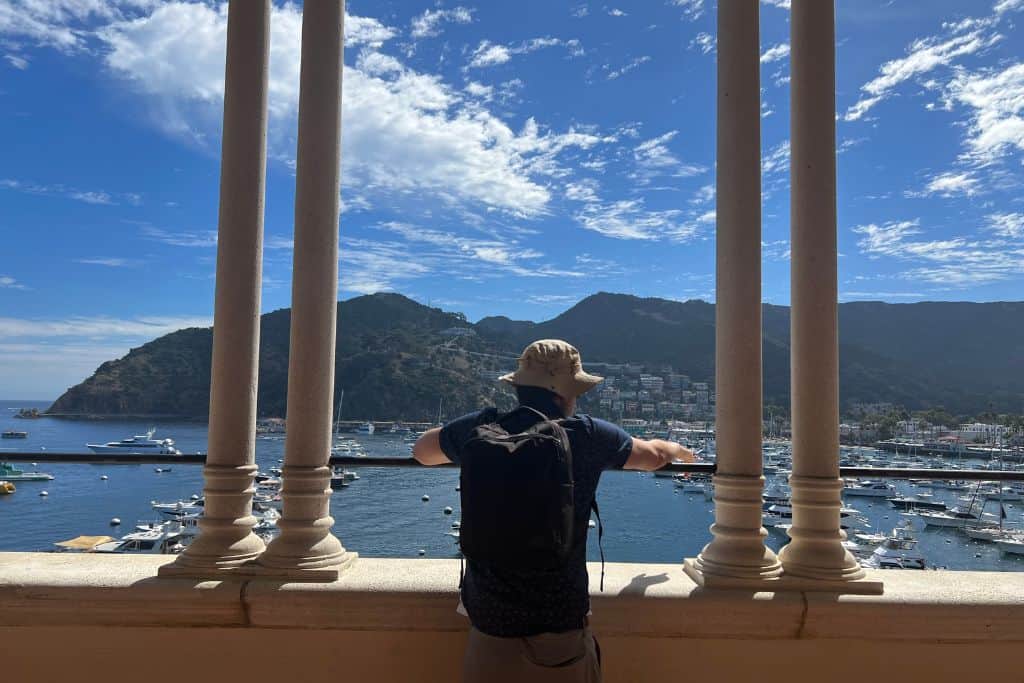 A man with a backpack on looks out onto the harbor and mountains of Catalina Island, California.