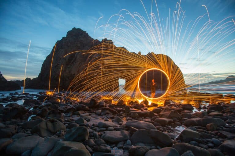 A large rock with a keyhole arch through it in the water next to a beach filled with rocks as someone stand with a fire work.