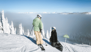 A woman standing on skies looking at a child knelt in the snow on a snowboard, both on a very snowy mountain.