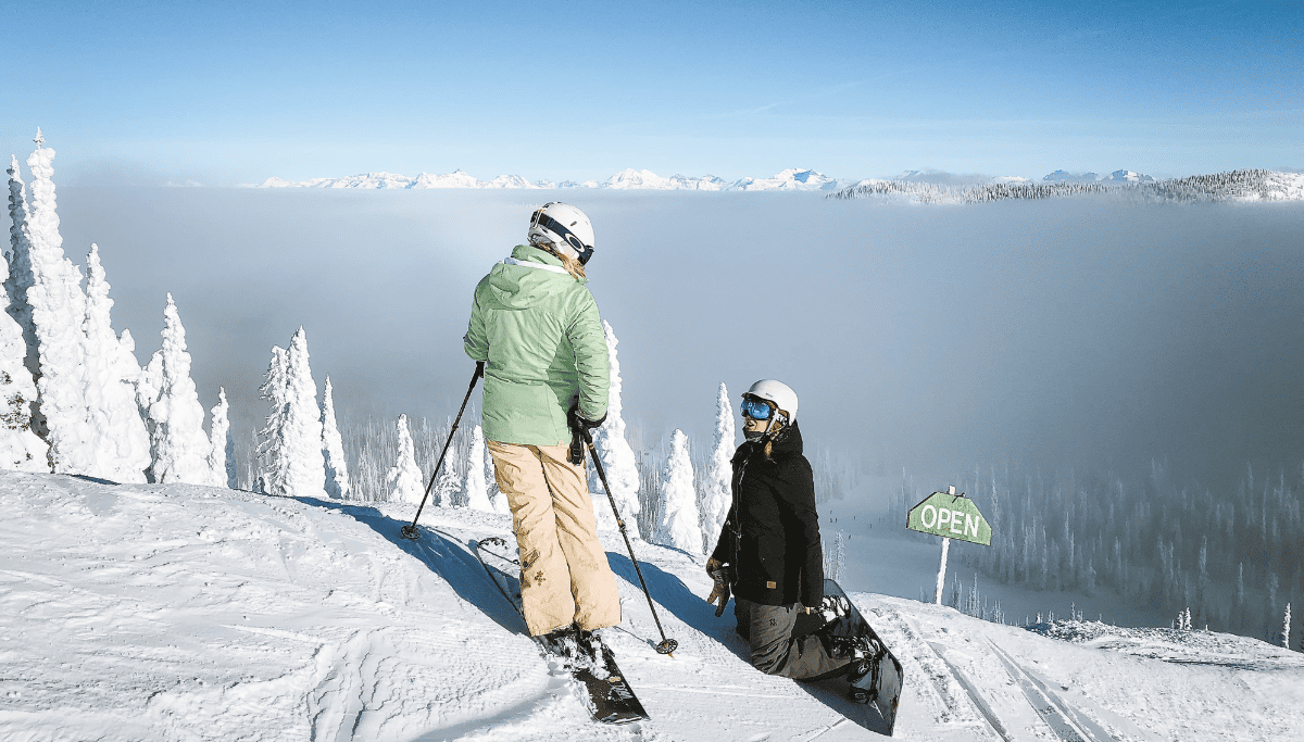 A woman standing on skies looking at a child knelt in the snow on a snowboard, both on a very snowy mountain.