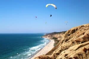 A beach at the bottom of a cliff with many parasailing people.