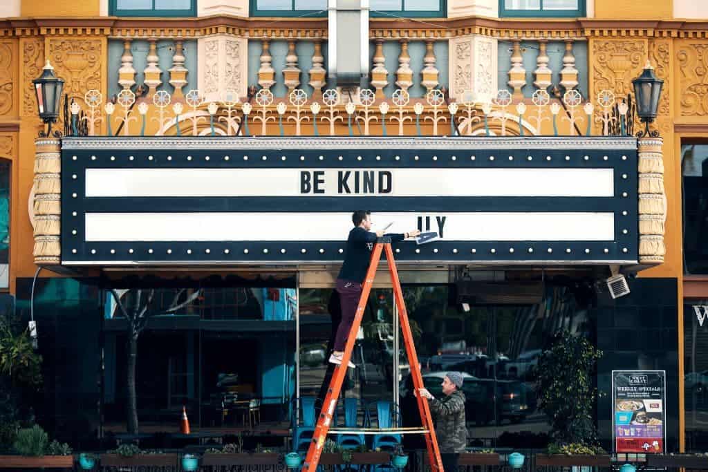 The marque in front of a theater that reads, "Be kind," A man on a ladder is taking down letters from the sign. 