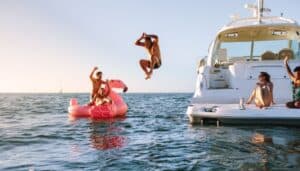 A person on a blow up swan and another person jumping in the water off a yacht while two women look on from the yacht deck.
