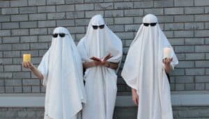Three people in sheets and sunglasses being ghosts. The middle one is looking down at a book. The ghost on the right is holding a candle.