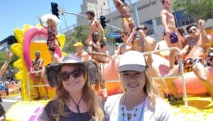 Two women smiling for the camera in front of a car full of half naked men during Gay Pride Parade in West Hollywood.