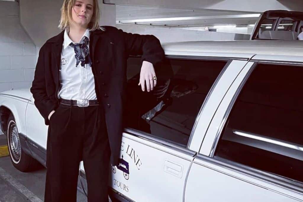 A woman standing next to a limo. She is wearing a white shirt and a black suit.
