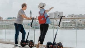 Two people next to Segways looking out onto water. The women is pointing at something toward the water and is wearing a bright red backpack.