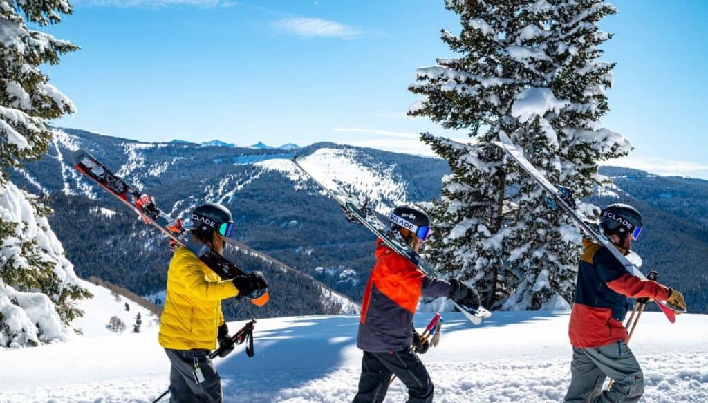 three people walking with skies on their shoulders across a snowy mountain. 