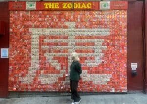 A girl stand infront of a red and white zodia sign in tiles on a giant wall.