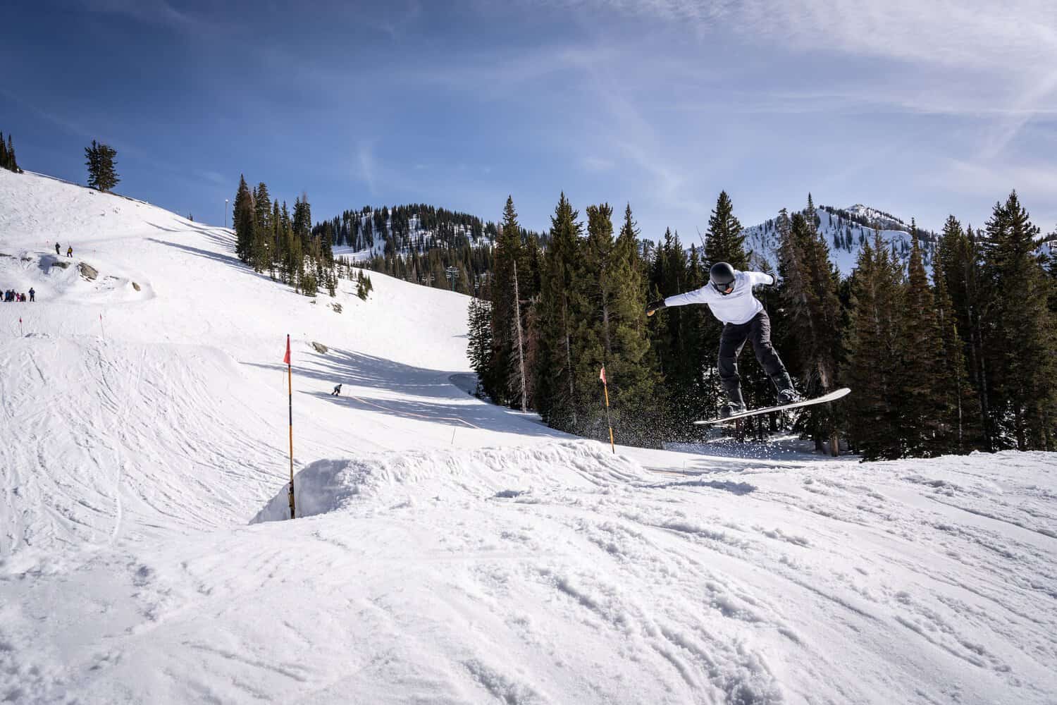 A snowboarder getting some air on a snowy mountain with large pine trees in the background.