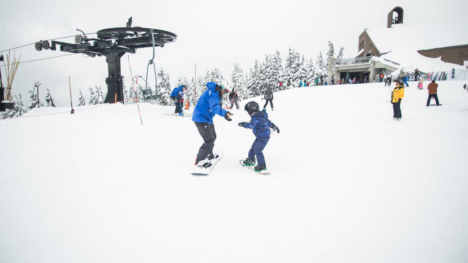 A man helping a child learn to snowboard on the top of a snowy mountain with a chair lift behind them.