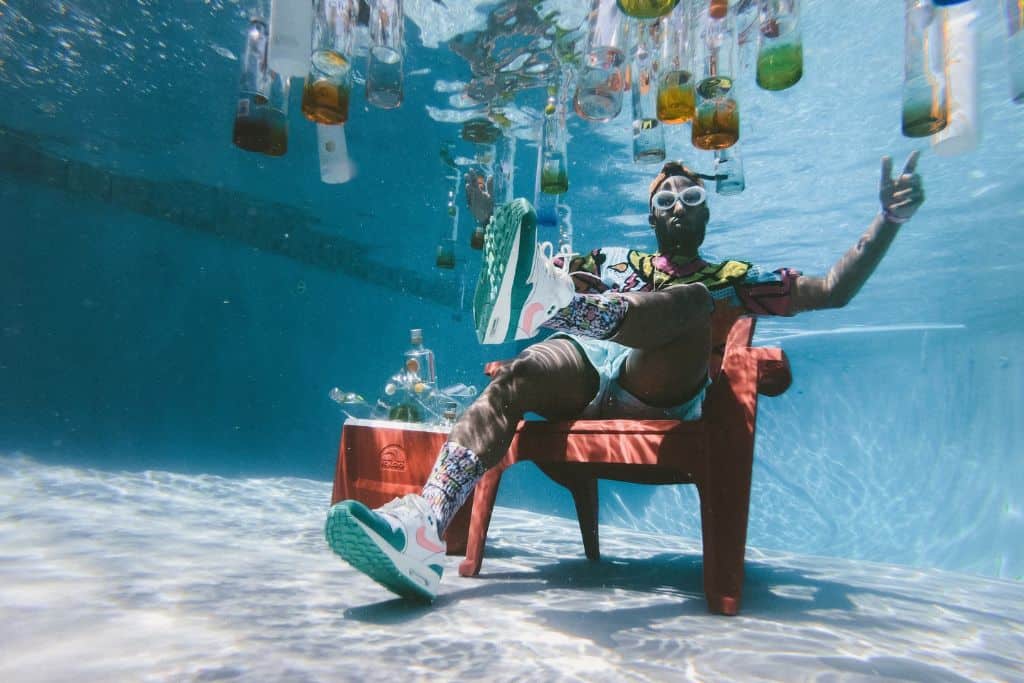 A man sitting in a chair at the bottom of a pool with glasses floating to the top. 
