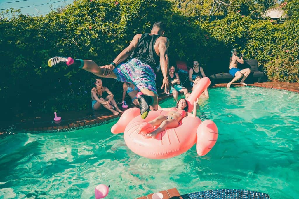 A man jumping into a pool onto a raft with people on the side of the pool laughing. 