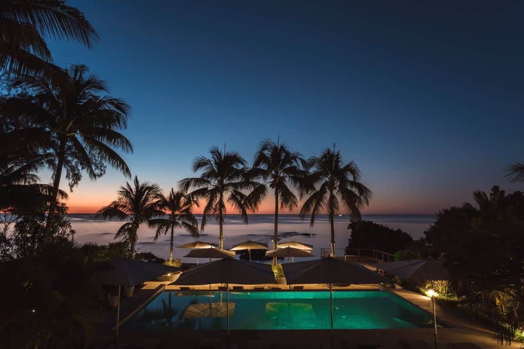 A pool next to the beach in the tropics with palm trees over the pool. 