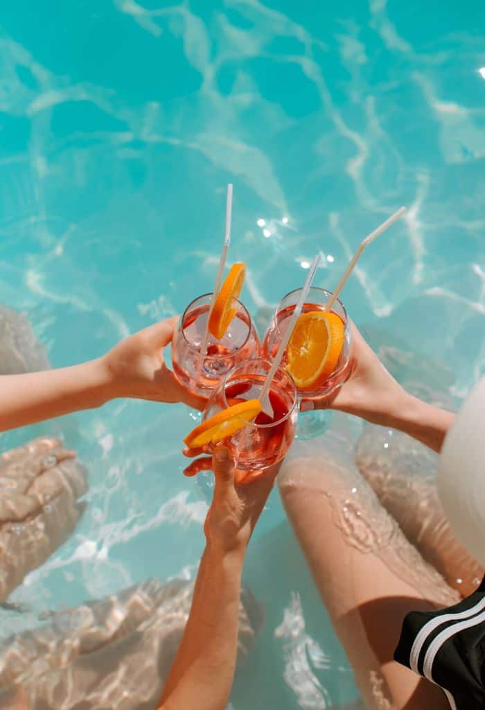 Three hands clinking glasses with straws, a slice of orange in them over a pool. 